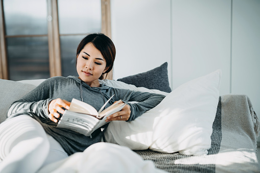 Relaxing young Asian woman lying in bed reading a book during the day. Beautiful sunlight illuminated her through window