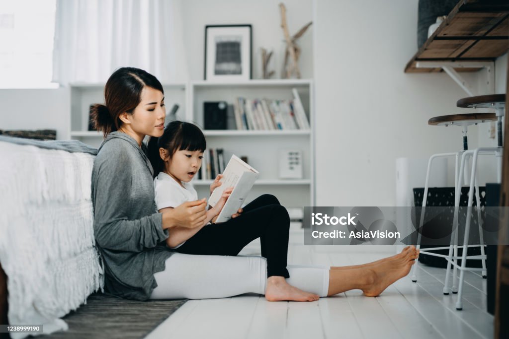 Young Asian mother sitting on the floor in the bedroom reading book to little daughter, enjoying family bonding time together at home Child Stock Photo