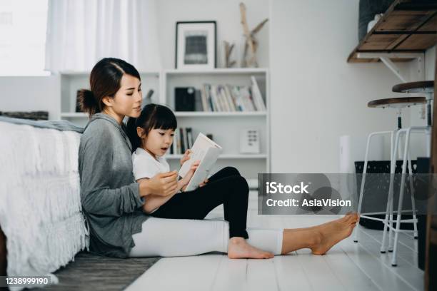 Young Asian Mother Sitting On The Floor In The Bedroom Reading Book To Little Daughter Enjoying Family Bonding Time Together At Home Stock Photo - Download Image Now