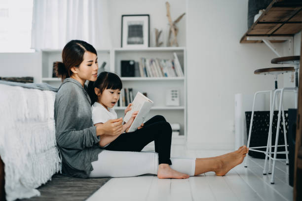 young asian mother sitting on the floor in the bedroom reading book to little daughter, enjoying family bonding time together at home - child reading mother book imagens e fotografias de stock