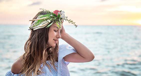 Beautiful young woman with a wreath of flowers on her head by the ocean at sunset.