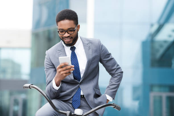 Handsome businessman texting on phone sitting on bike Way To Office. Smiling african manager in specs texting on phone, sitting on bicycle outdoors near office, free space afro man stock pictures, royalty-free photos & images