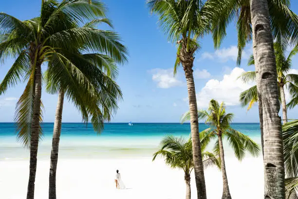 (Selective focus) Stunning view of woman walking on a white sand beach bathed by a turquoise sea, beautiful coconut palm trees in the foreground. White Beach, Boracay Island, Philippines.