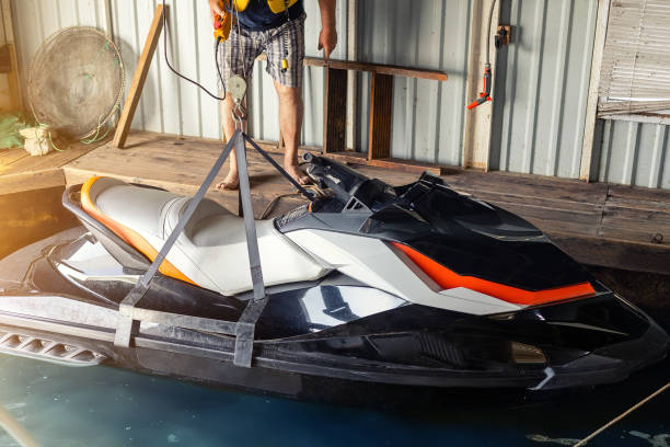 istock Senior man launching or lifting modern fast jetski hanged on strpas with wire hoist rail crane for dry storage. Boat garage interior for boats and jet-ski storage, service and maintenance 1239229580