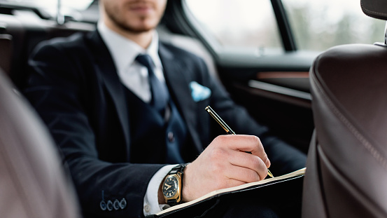 Unrecognizable businessman writing in notebook with pen sitting in back seat of luxury car, selective focus