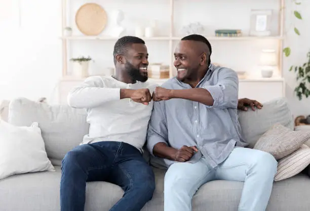 Father-Son Relationships Concept. Adult Black Man And His Senior Father Bumping Fists While Sitting On Couch At Home