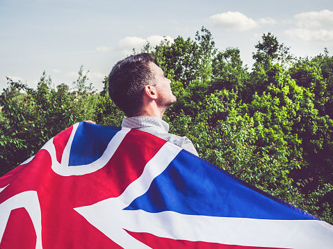Attractive man holding British Flag on blue sky background on a clear, sunny day. View from the back, close-up. National holiday concept
