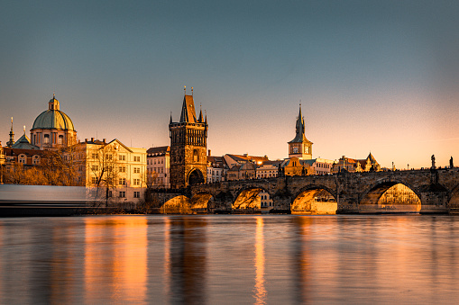 Panoramic highly detailed view from distance towards Vltava river and the famous Charles bridge in beautiful City of Prague, the capital of the Czech republic. The image is a long exposure with blurred passing boat and constant tourist crowds passing the iconic bridge sunlit by the setting Sun during afternoon on a clear day. Shot on Canon EOS R system with premium lens for highest quality.