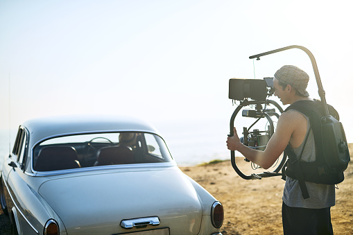 Shot of a focused young man shooting a scene with people in a car with a state of the art video camera outside during the day