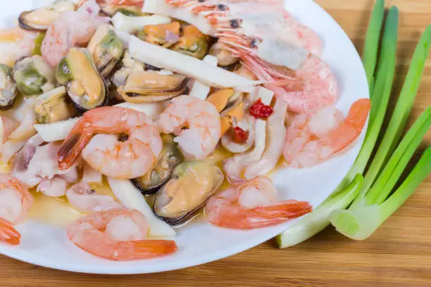 Various cooked seafoods - shrimps, prawn tails, mussels peeled from shells, calamari slices on the white dish, green onion on the wooden surface, fragment close-up