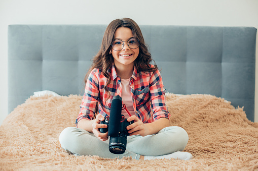 Gladsome cute teenage girl wearing glasses sitting on the bed and smiling while having a camera with microphone in her hands