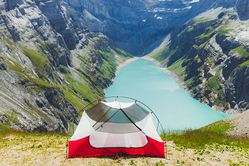 Woman hiker and waking up from the red tent camping on the top of the mountains with scenic view of the turquoise colored lake and mountain peaks in Swiss Alps
