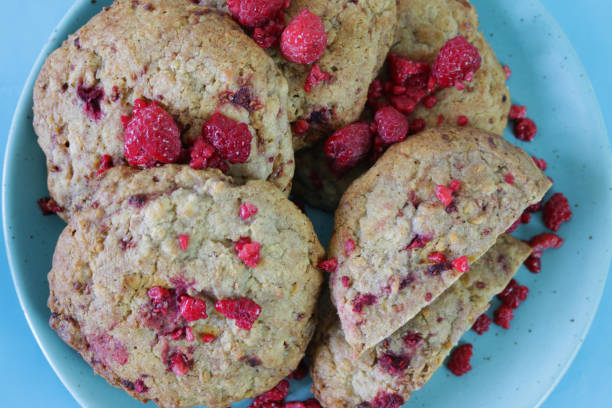 Stock photo showing elevated view of heap of freshly baked white chocolate chip raspberry cookies on a blue ceramic plate with fresh raspberry sprinkled on top with blue background. New York style Levine homemade white chocolate raspberry cookie / biscuits in kitchen bakery. imagen de galletas de frambuesa de chocolate blanco recién horneadas sobre un plato de cerámica azul sobre frambuesa fresca espolvoreada sobre fondo azul, montón de galletas / galletas de frambuesa de chocolate blanco casero al estilo neoyorquino, vista