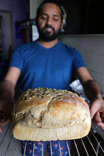 Stock photo showing Indian man with beard holding fresh homemade artisan bread loaf on cooling rack, golden brown, fresh from oven, crusty granary bread topped with sunflower seeds, closeup