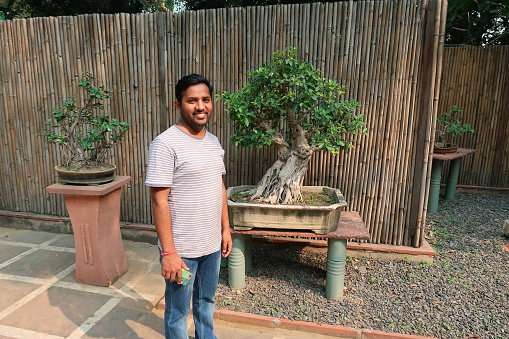 Stock photo of handsome Indian man wearing stripped t-shirt and denim jeans standing in the middle of paved courtyard in garden featuring Japanese elements of bonsai trees on plinths displays