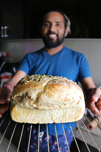 Stock photo showing Indian man with beard holding fresh homemade artisan bread loaf on cooling rack, golden brown, fresh from oven, crusty granary bread topped with sunflower seeds, wearing blue T-shirt