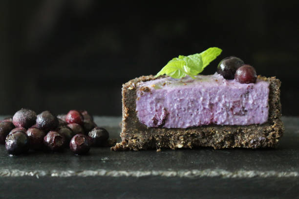 Stock photo showing half a miniature blueberry cheesecake with buttery biscuit base, blueberry mascarpone cream cheese for dessert pudding. imagen de tarta de queso de arándano en miniatura con base de galletas mantecosas sobre fondo oscuro rodeado de bayas, medio pudín de postre de queso crema de mascarpone de arándano cubierto con arándanos frescos y guarnición de hoja de menta