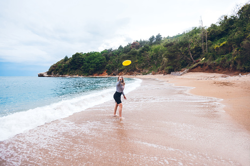 Young Caucasian woman playing with frisbee on sea shore.