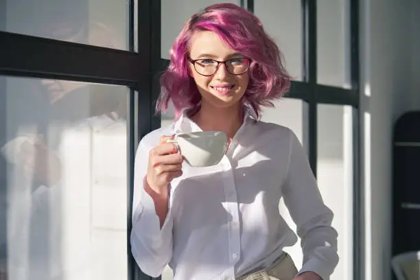 Smiling young adult hipster teen girl pink hair wearing white shirt and glasses holding tea cup looking at camera standing at glass wall in modern sunny home office. Head shot close up portrait.
