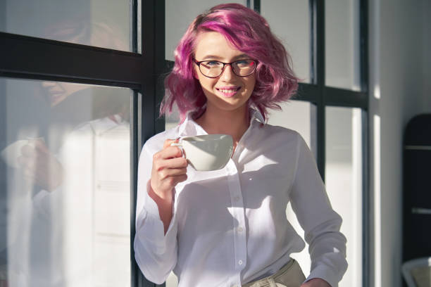улыбаясь молодой взрослый хипстер подросток девушка розовые волосы в белой рубашке и очках проведения чашку чая глядя на камеру, стоящую н� - gen z стоковые фото и изображения