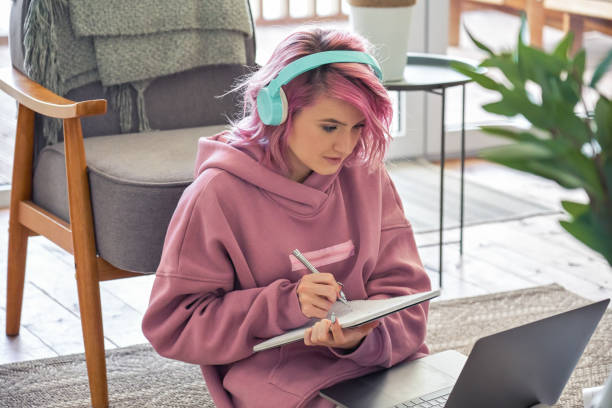 focalizzato hipster teen girl studentessa universitaria rosa capelli indossare cuffie scrivere note guardando webinar videoconferenza online chiamata su computer portatile sedersi sul pavimento di apprendimento di lavoro online a casa. - girl casual foto e immagini stock