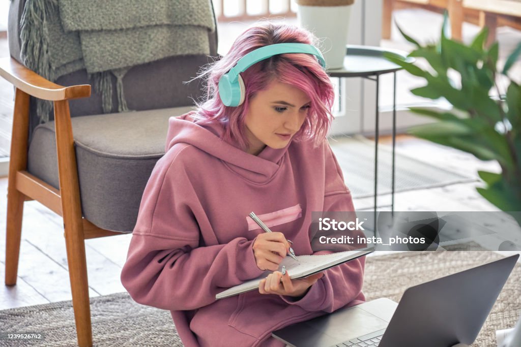 Focused hipster teen girl school college student pink hair wear headphones write notes watching webinar online video conference calling on laptop computer sit on floor working learning online at home. Teenager Stock Photo