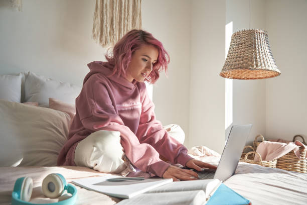 хипстер подросток школьница с розовыми волосами носить балахон с помощью ноутбука сидя в постели расстояние elearning онлайн учебный курс для � - gen z стоковые фото и изображения