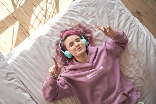 Feliz chica adolescente divertida con el pelo rosa llevan auriculares acostados en la cama cómoda escuchando nueva música pop disfrutando de la canción cantando con los ojos cerrados relajándose en el acogedor dormitorio en casa. Vista superior desde a photo