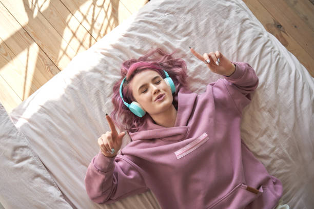 het gelukkige grappige tienermeisje met roze haar draagt hoofdtelefoon die in comfortabel bed ligt dat nieuwe popmuziek luistert die van het zingen lied met gesloten ogen geniet ontspannend in gezellige slaapkamer thuis. bovenste uitzicht van bovenaf. - music stockfoto's en -beelden
