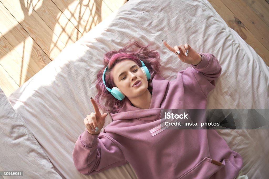 Happy funny teen girl with pink hair wear headphones lying in comfortable bed listening new pop music enjoying singing song with eyes closed relaxing in cozy bedroom at home. Top view from above. Teenager Stock Photo