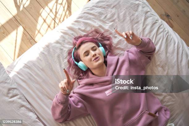 Happy Funny Teen Girl With Pink Hair Wear Headphones Lying In Comfortable Bed Listening New Pop Music Enjoying Singing Song With Eyes Closed Relaxing In Cozy Bedroom At Home Top View From Above Stock Photo - Download Image Now