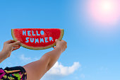 The sun shines. A piece of watermelon against a blue sky. Children's hands are holding a slice of watermelon with the text Hello Summer. Summer time concept