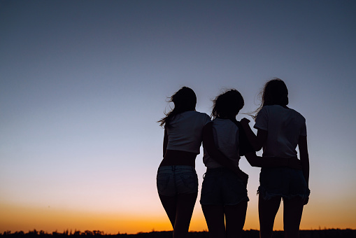 Silhouette of three young girls standing on the beach and looking to a sunset. Summer holidays, vacation, relax and lifestyle concept.