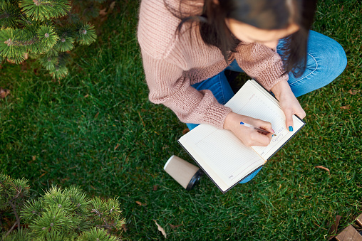 Top view of calm and concentrated female student planning her day in a paper notepad on a green grass lawn outside