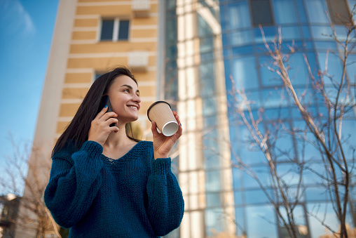 Nice cheerful lady looking aside with a shiny smile holding cup of hot drink in hand and listening to someone by her smartphone