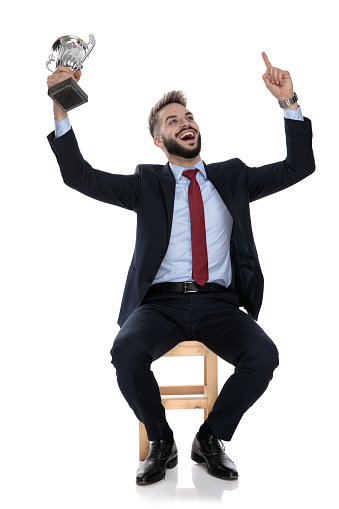 excited young businessman holding trophy,celebrating victory with hands in the air, sitting isolated on white background