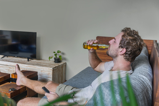Close up shot of lazy Deutsch man relaxing at home , drinking beer while watching tv alone, during Covid 19 lockdown