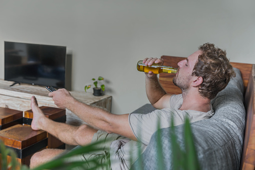 Close up shot of lazy Deutsch man drinking beer while changing tv channels at home, during Coronavirus lockdown