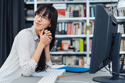 Pretty young businesswoman sitting thinking deeply at a desktop computer in the office looking back over her shoulder with a pensive expression