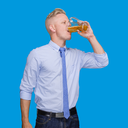 Front view of with blond hair young male manual worker standing in front of blue background wearing smart casual who is drinking and showing hand raised and holding craft beer