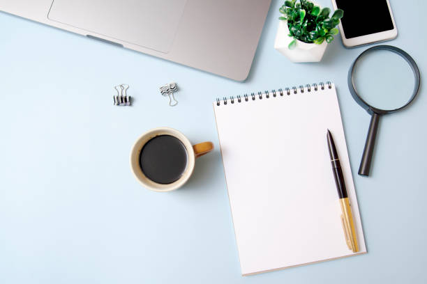 istock Top view workspace with a notebook (mock-up), magnifier, coffee, laptop on a blue background. Desktop business concept 1239803555