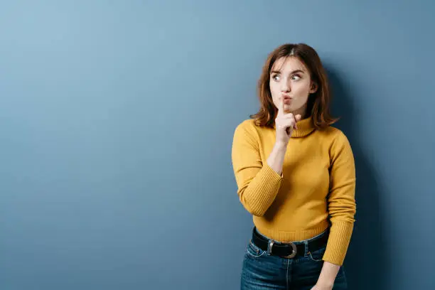 Young woman making a shushing gesture with her finger to her lips as she asks for silence or secrecy as she glances to the side over a blue studio background with copy space