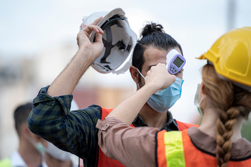 Factory woman worker in a face medical mask and safety dress used measures temperature at worker people standing on queue with a non-contact infrared thermometer.
