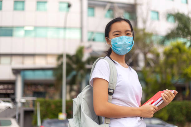바이러스 보호: 보호 얼굴 마스크를 착용하고 카메라를 보고 있는 아시아 여학생의 야외 초상화 - student printed media walking clothing 뉴스 사진 이미지