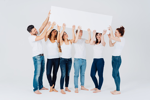 Group of happy men and women standing barefoot in jeans and white tops holding up a blank white banner sign or placard with copy space on a white studio background