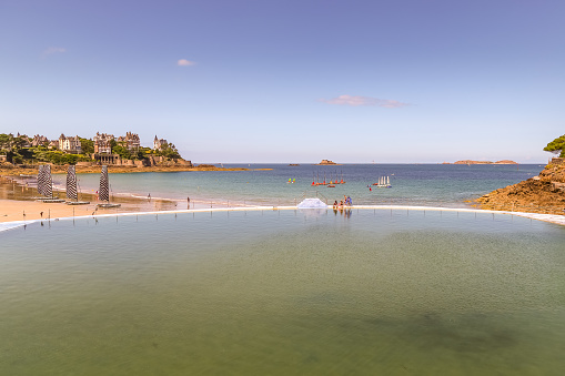 Dinard, Ille-et-Vilaine, France - July 31 2018: Piscine de mer pool, people sit on the waters edge at lowtide with a view directly across the harbour. Small sailboats surround the harbour in the afternoon sun.