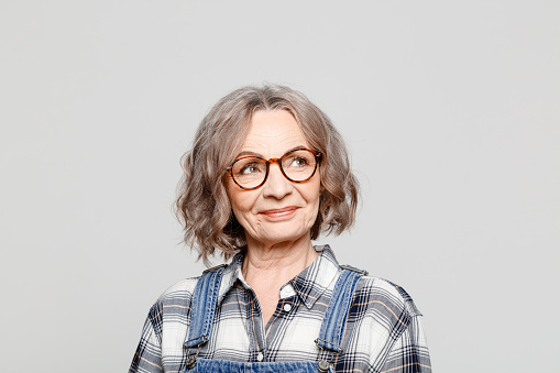 Headshot of elderly lady wearing dungarees and checkered shirt standing against grey background, peeking at copy space. Studio shot of female designer.