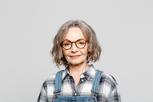 Headshot of elderly lady wearing dungarees and checkered shirt standing against grey background, smiling at camera. Studio shot of female designer.