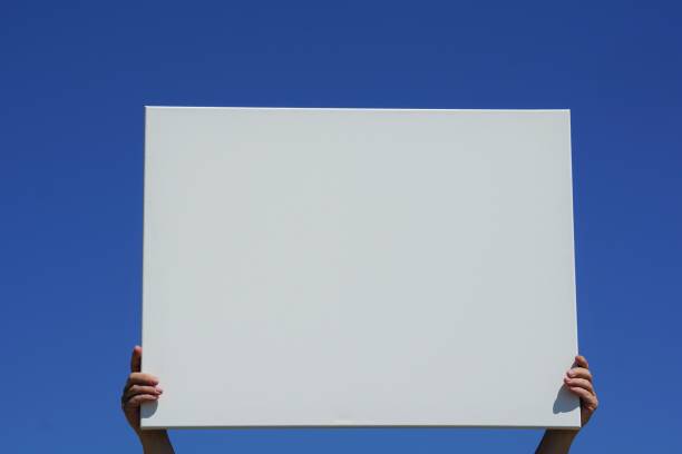 istock woman holds a blank white board in her hands. Blue sky in background 1240090807