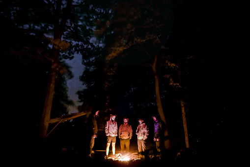 Group of men, backpackers, tourist standing around campfire in forest under oak trees and sky is visible between trees in White Carpathians in Czechia (Carpathian mountain range)